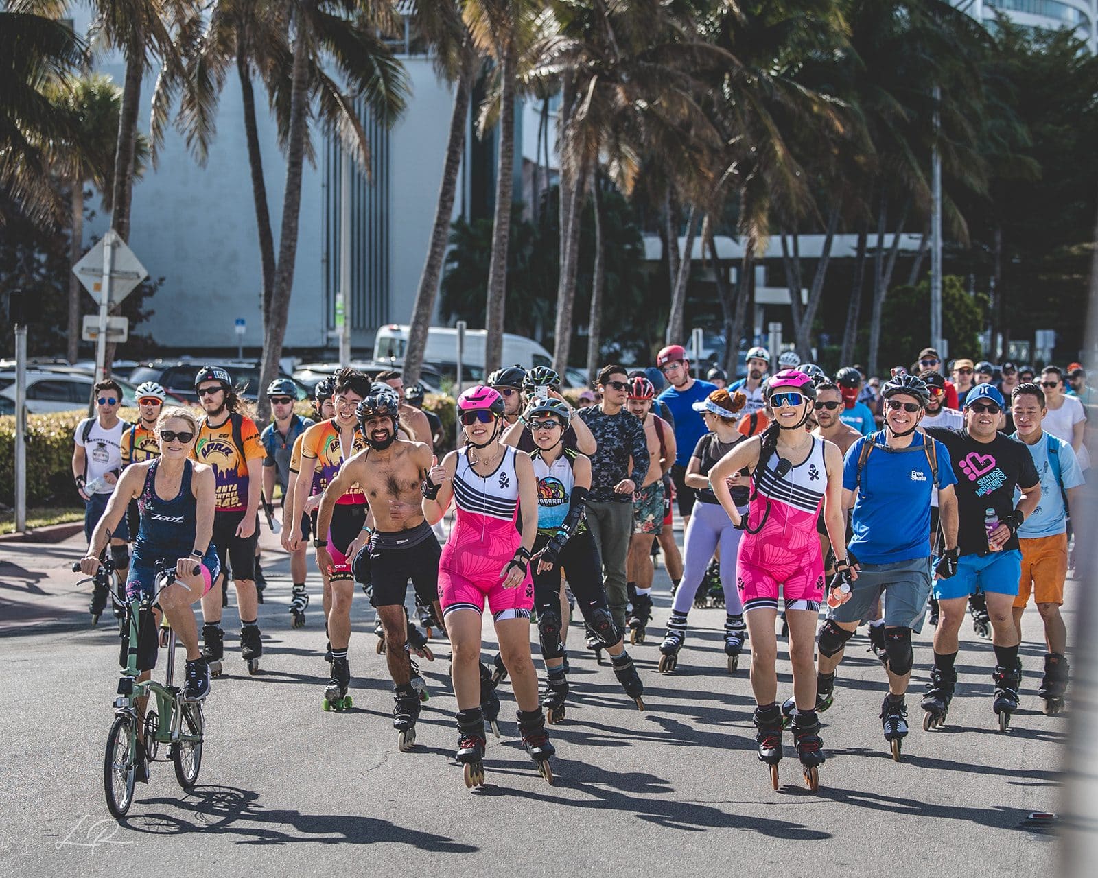 A crowd of skaters in bright kits rolling down a sunny Miami street